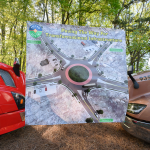 A banner featuring a rendering of the proposed 5-way roundabout suspended between the noses of a red vehicle and a brown vehicle.